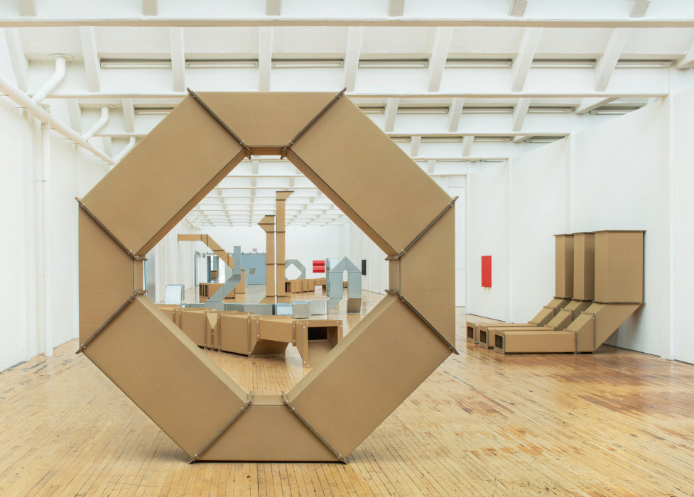 Installation of cardboard and tin-looking sculptures in large room with white walls and a wood floor. Many sculptures are visible through a wheel shaped cardboard sculpture in the foreground.